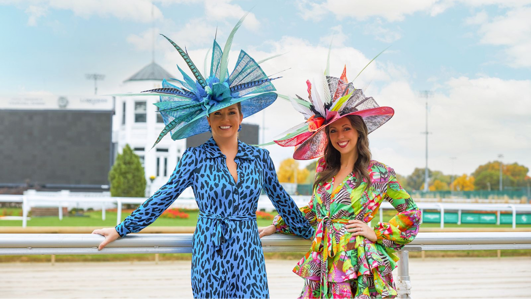 Two women in colorful wide-brim derby hats with tall feather trims pose together at a racecourse rail.