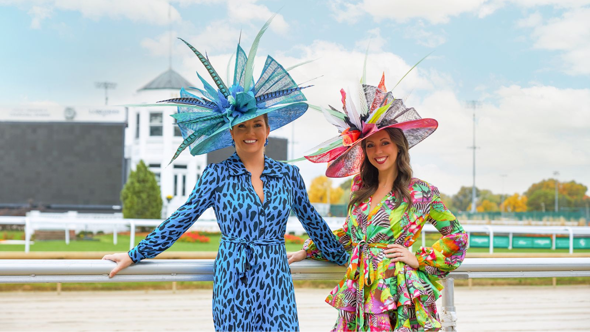 Two women in colorful wide-brim derby hats with tall feather trims pose together at a racecourse rail.