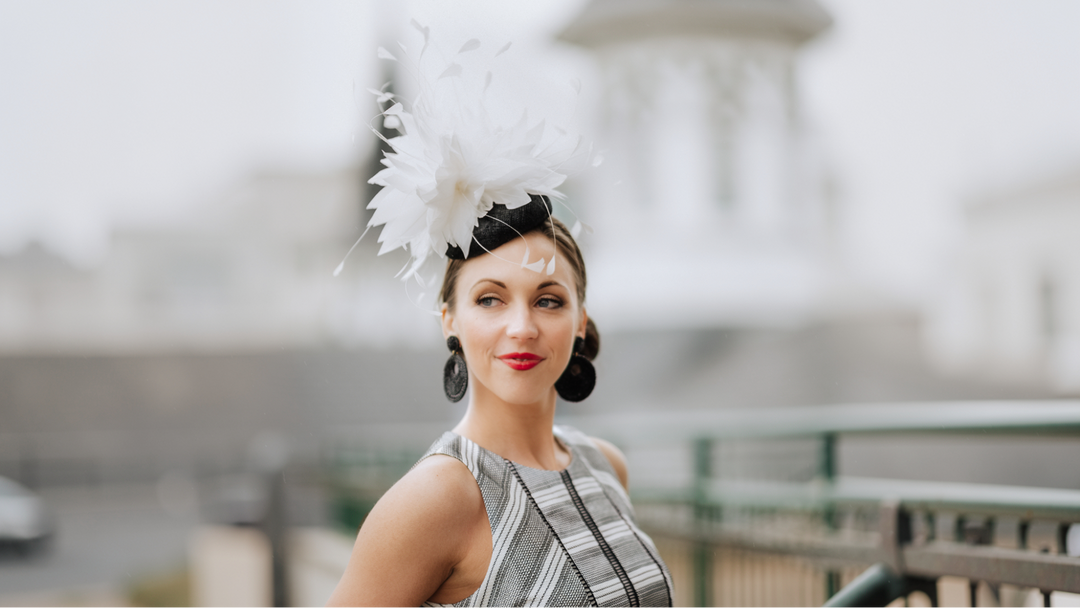 Woman wearing an elegant white feather fascinator with a striped dress, standing on a bridge near a domed building.