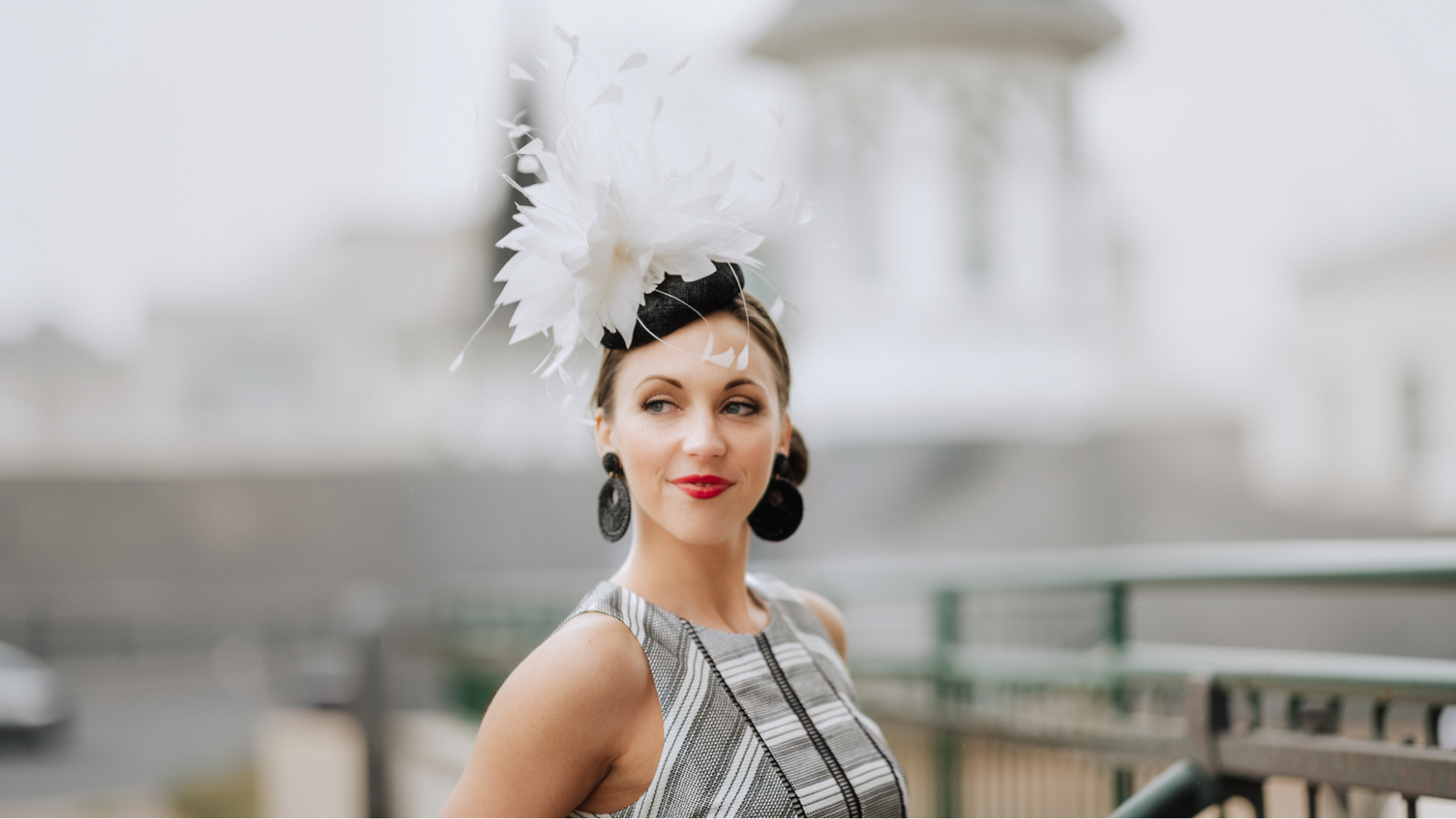 Woman wearing an elegant white feather fascinator with a striped dress, standing on a bridge near a domed building.