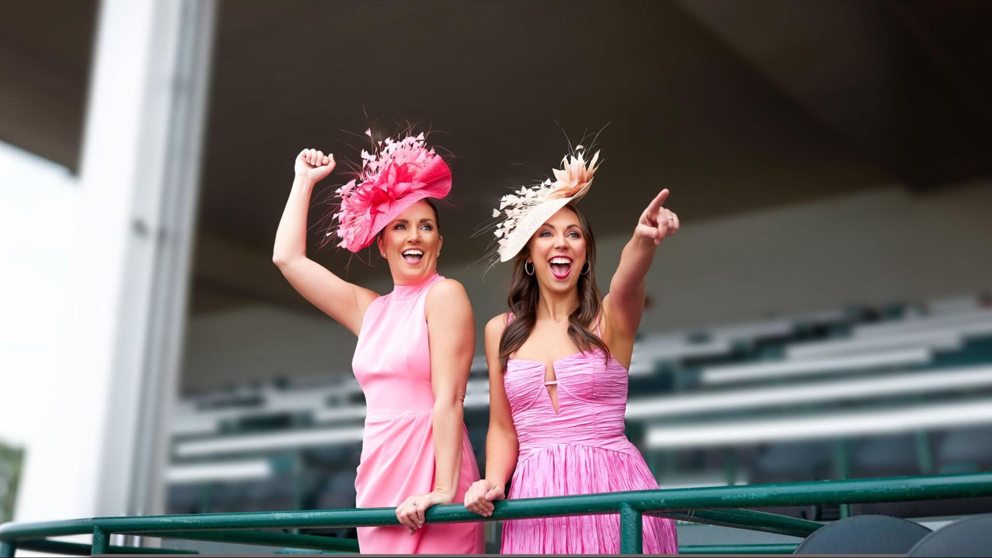 Two women in pink dresses wearing colorful fascinators cheering at Kentucky Derby grandstand