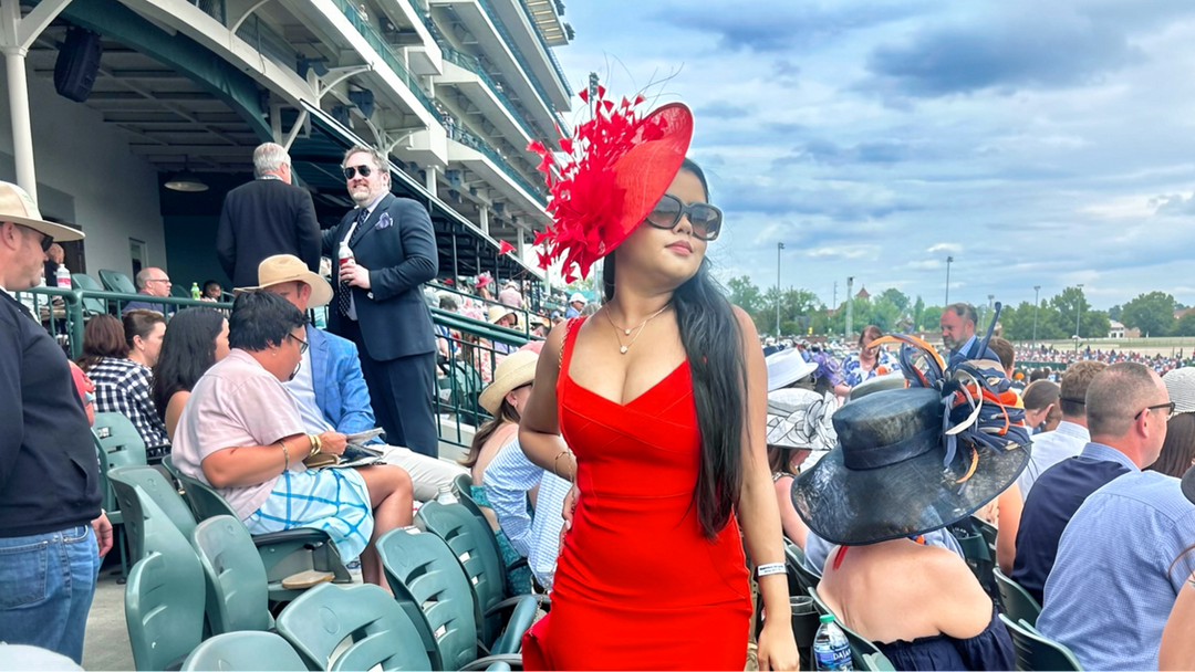 Woman in a red dress wearing a large red feather fascinator at a horse race, surrounded by spectators.