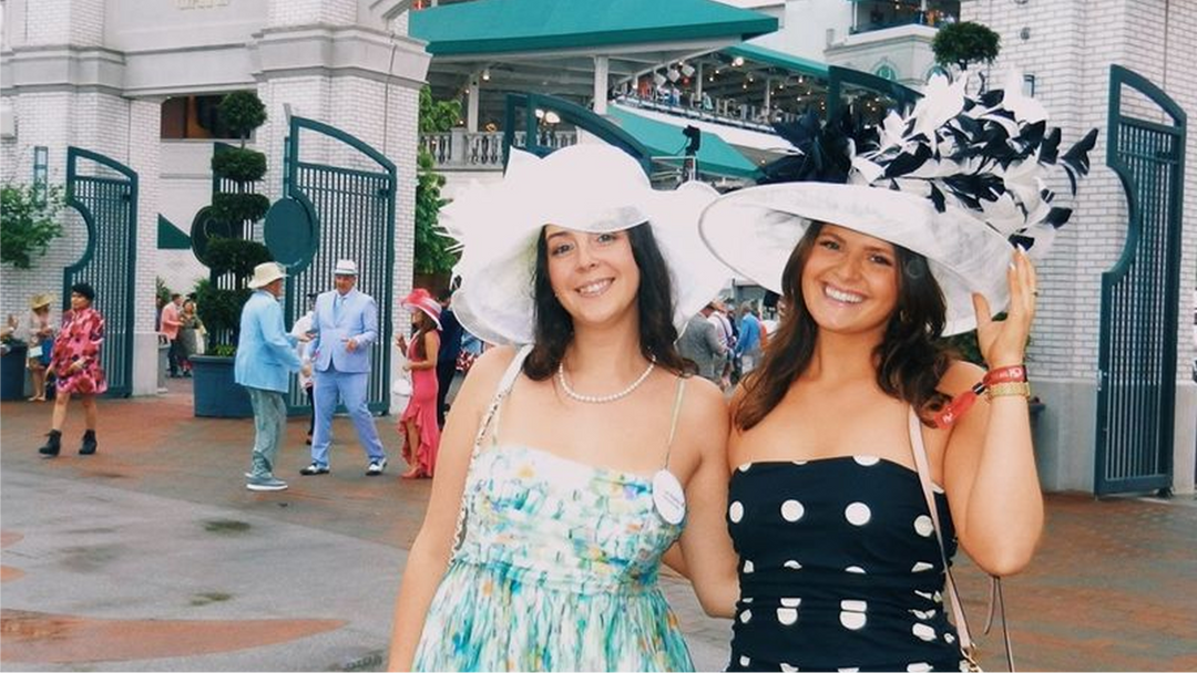Two smiling women wearing wide-brim white hats with black feather trim at an outdoor event entrance gate.
