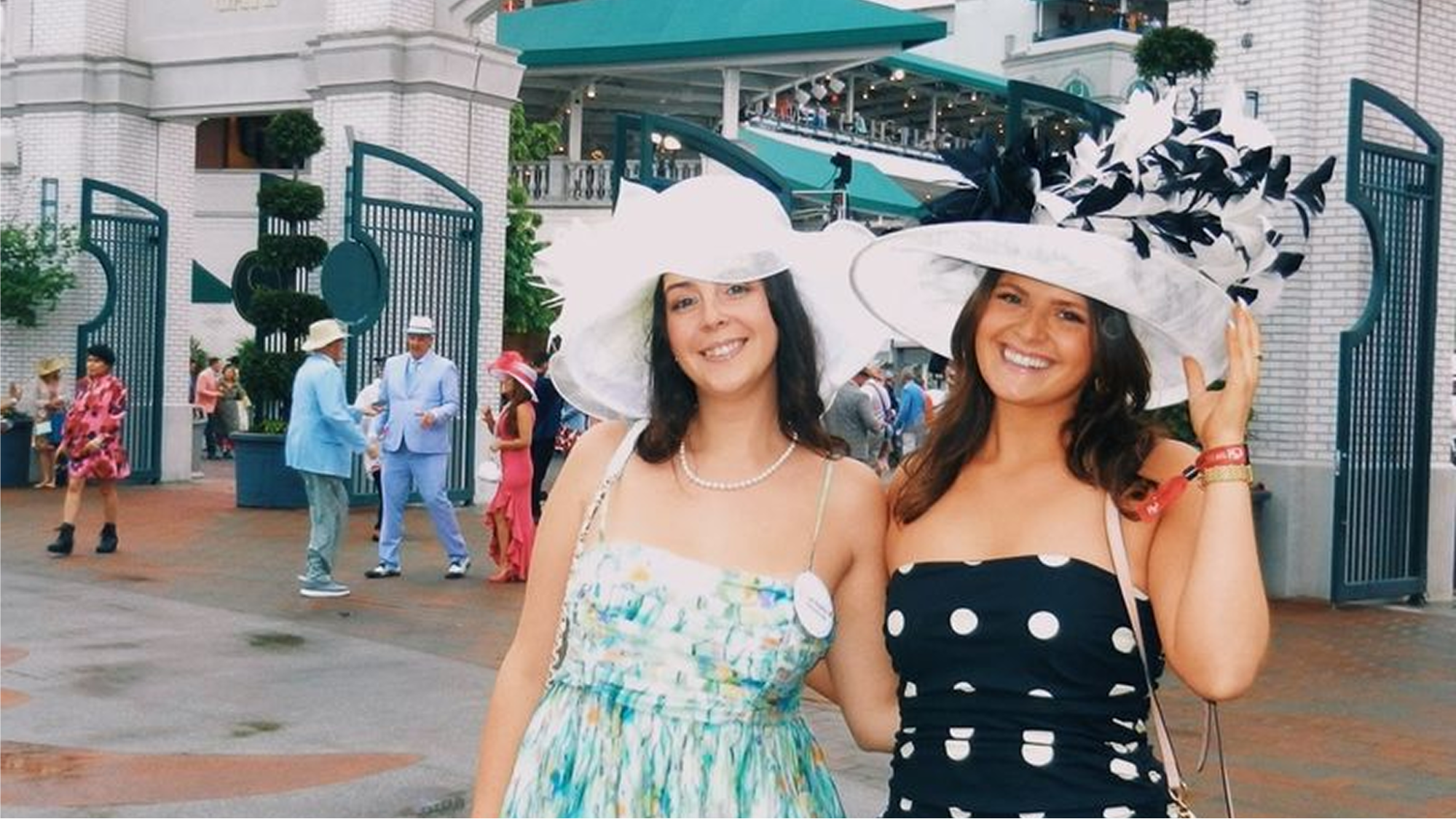 Two smiling women wearing wide-brim white hats with black feather trim at an outdoor event entrance gate.